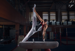 Male gymnast performing a routine on a pommel horse in a dimly lit gym, dressed in black and white attire with one leg extended upward.