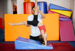 A child practices a handstand with the assistance of an adult in a gym. Colorful gym mats are stacked in the background.