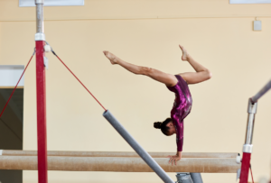A gymnast in a maroon leotard performs a handstand on a balance beam in a gymnasium.