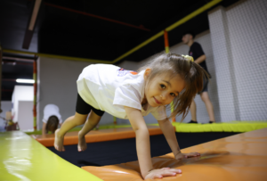 A young girl plays on a trampoline indoors, supporting herself with her hands. She's wearing a white shirt and black shorts. Another child and an adult are visible in the background.